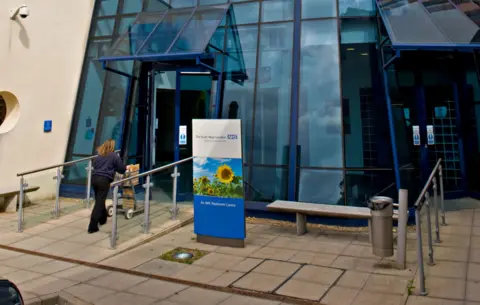 Alamy A sign outside the entrance to the South West London Orthopedic Centre, a glass building with the NHS logo on the front, where a staff member walks up to the door pushing a trolley, taken in Epsom in 2009.